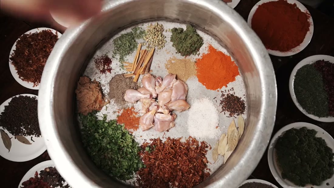 Overhead view of a steel vessel with twenty-odd spices arranged in segments around raw chicken