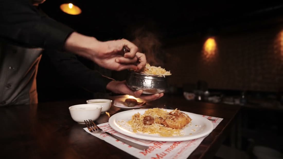 A server spooning biryani from a small handi onto a plate by candlelight